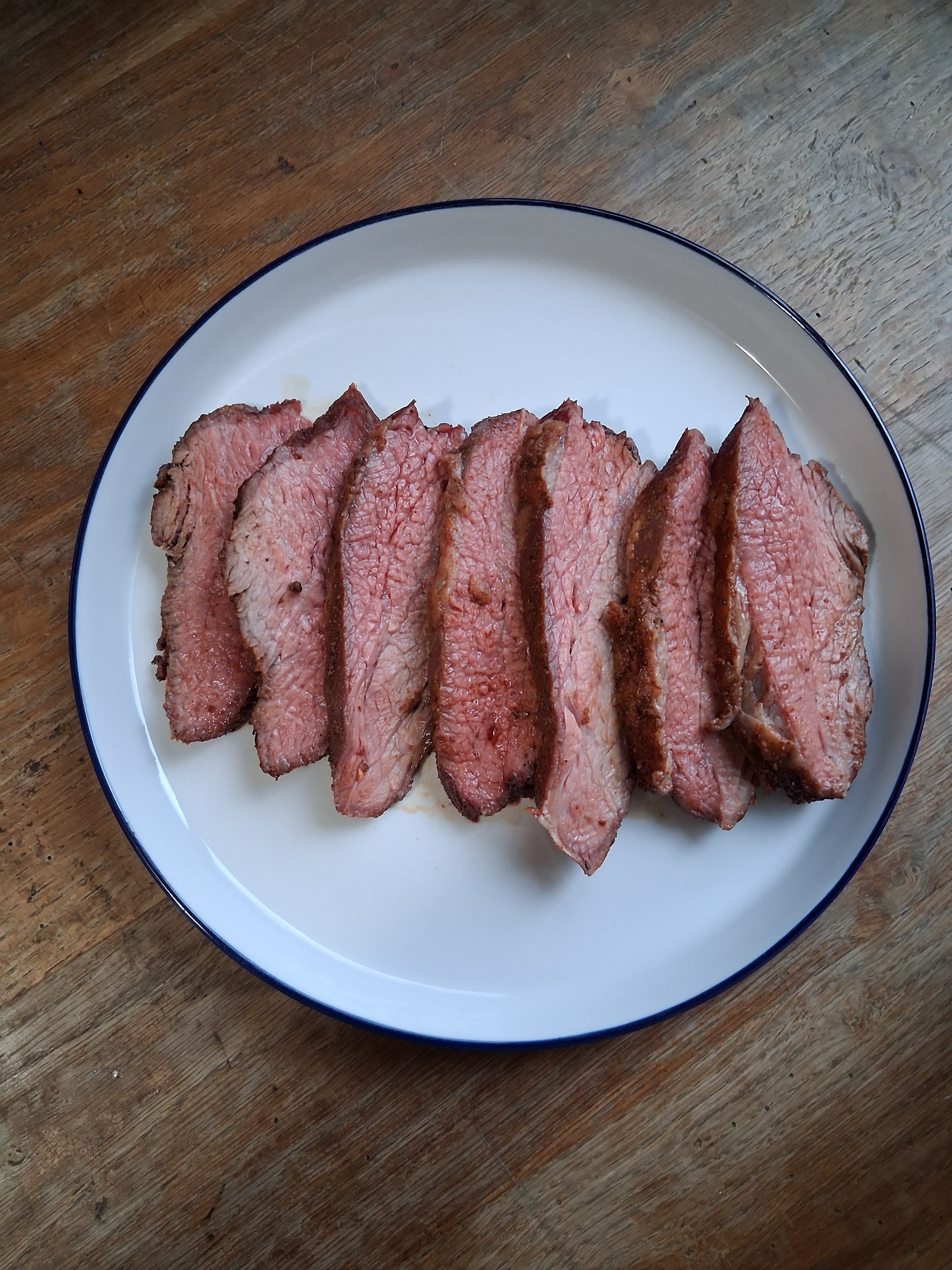Sliced medium-rare BBQ tri tip on a white plate on a wooden table, seasoned with Lucky Cajun Black Label and served with homemade Dr Pepper barbeque sauce.