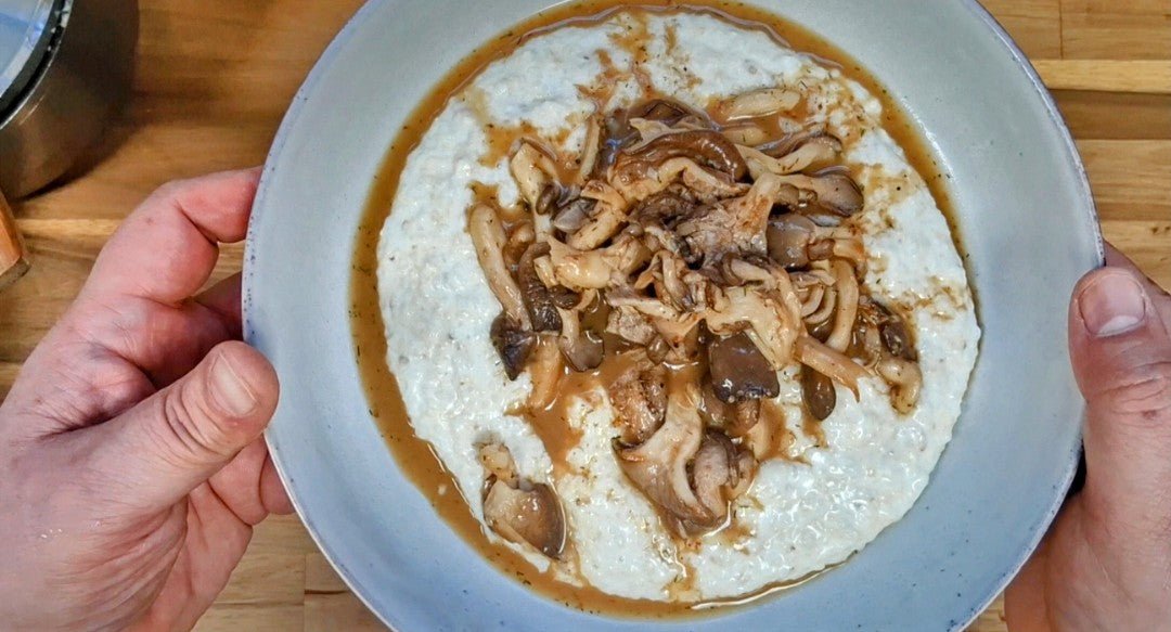 Chef Blackwell Smith holding a large white bowl of creamy stone-ground grits topped with glazed oyster mushrooms seasoned with Lucky Cajun Lucky Lemon Dilly Pepper fresh ground seasoning over a wooden cutting board