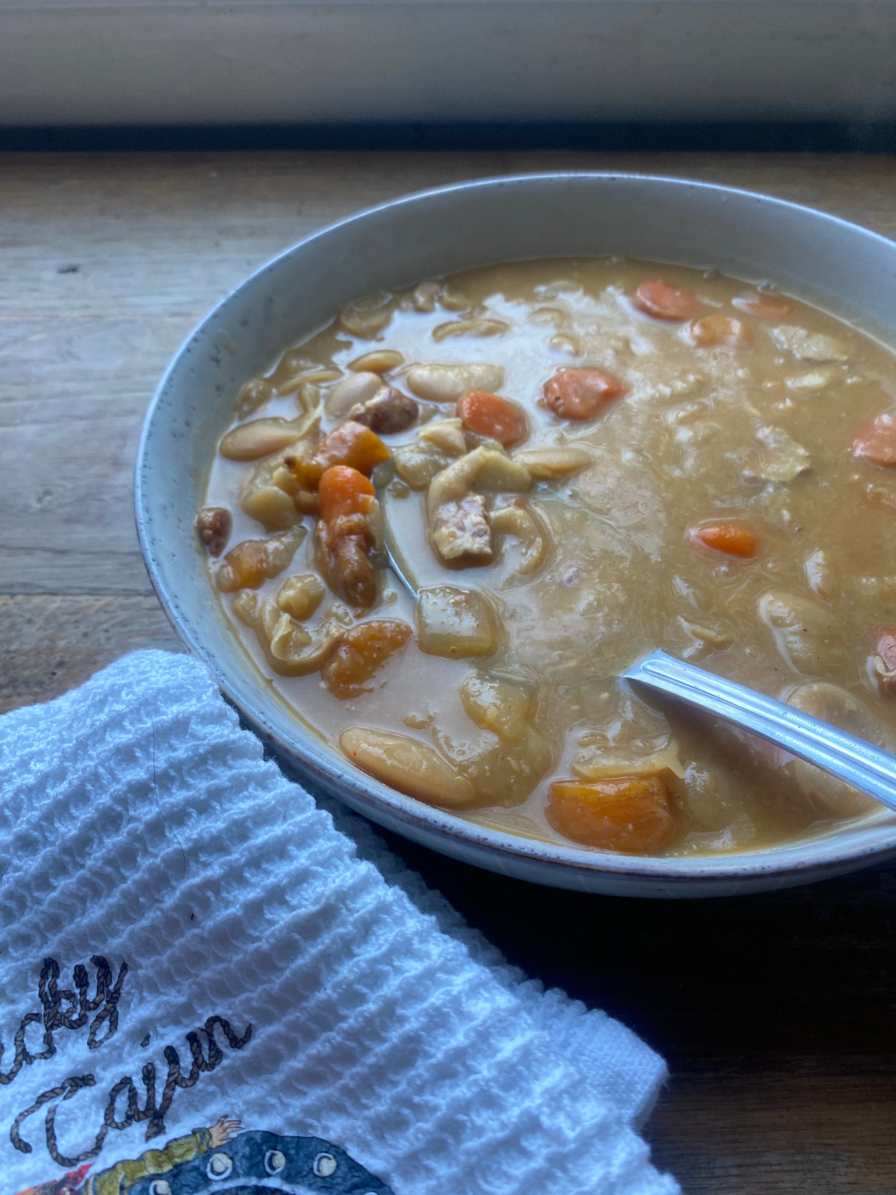 Close-up of hearty lima bean soup in a bowl with a spoon on a wooden table, made with Lucky Cajun Black Label seasoning and bacon.