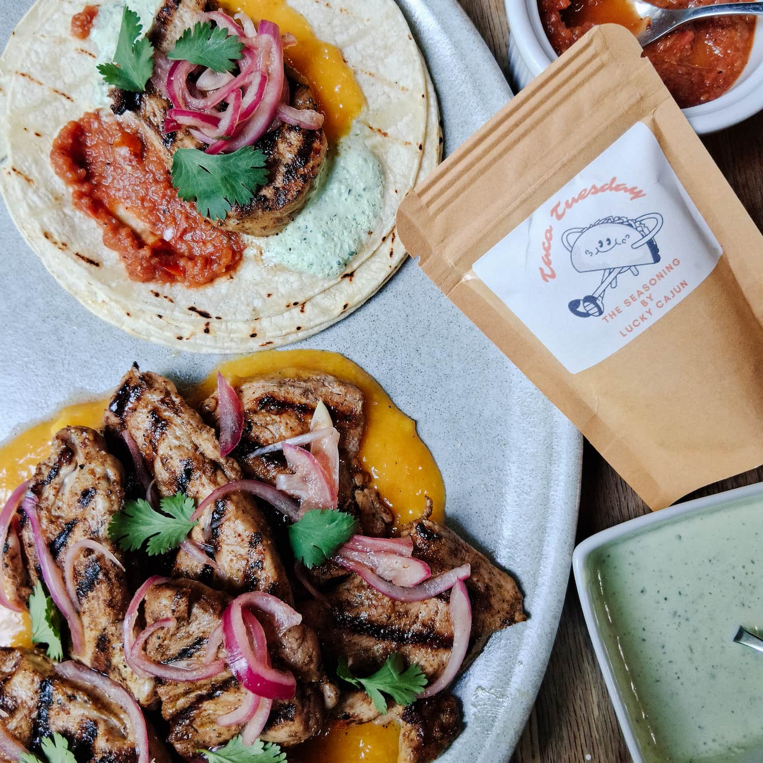 Lucky Cajun Taco Tuesday seasoning product photo alongside assembled pork tacos with roasted tomato salsa avocado jalapeño cream mango sauce and grilled pork tenderloin slices