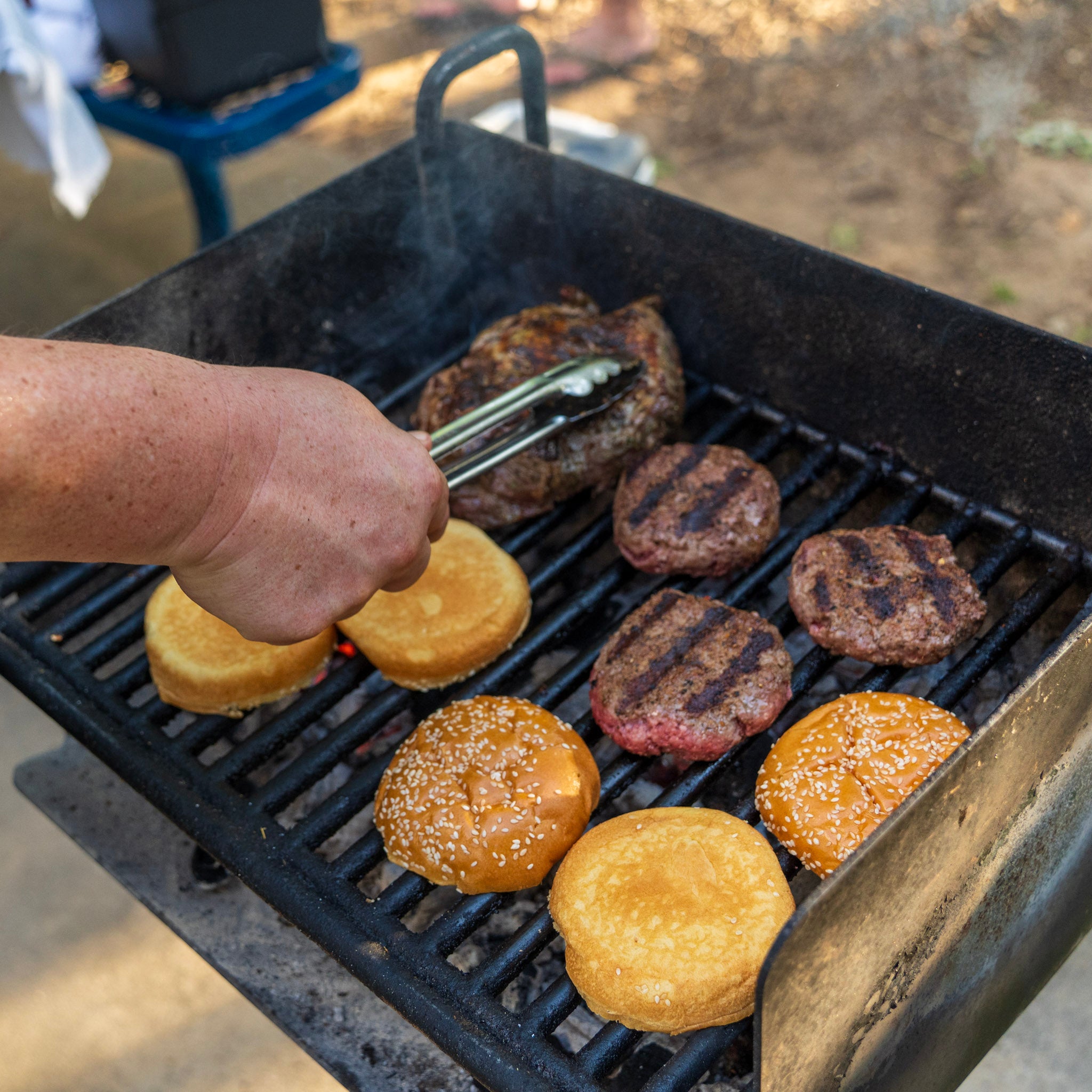 Chef Blackwell Smith's hands holding tongs grilling Daytona Sizzle burgers with buns on the grill