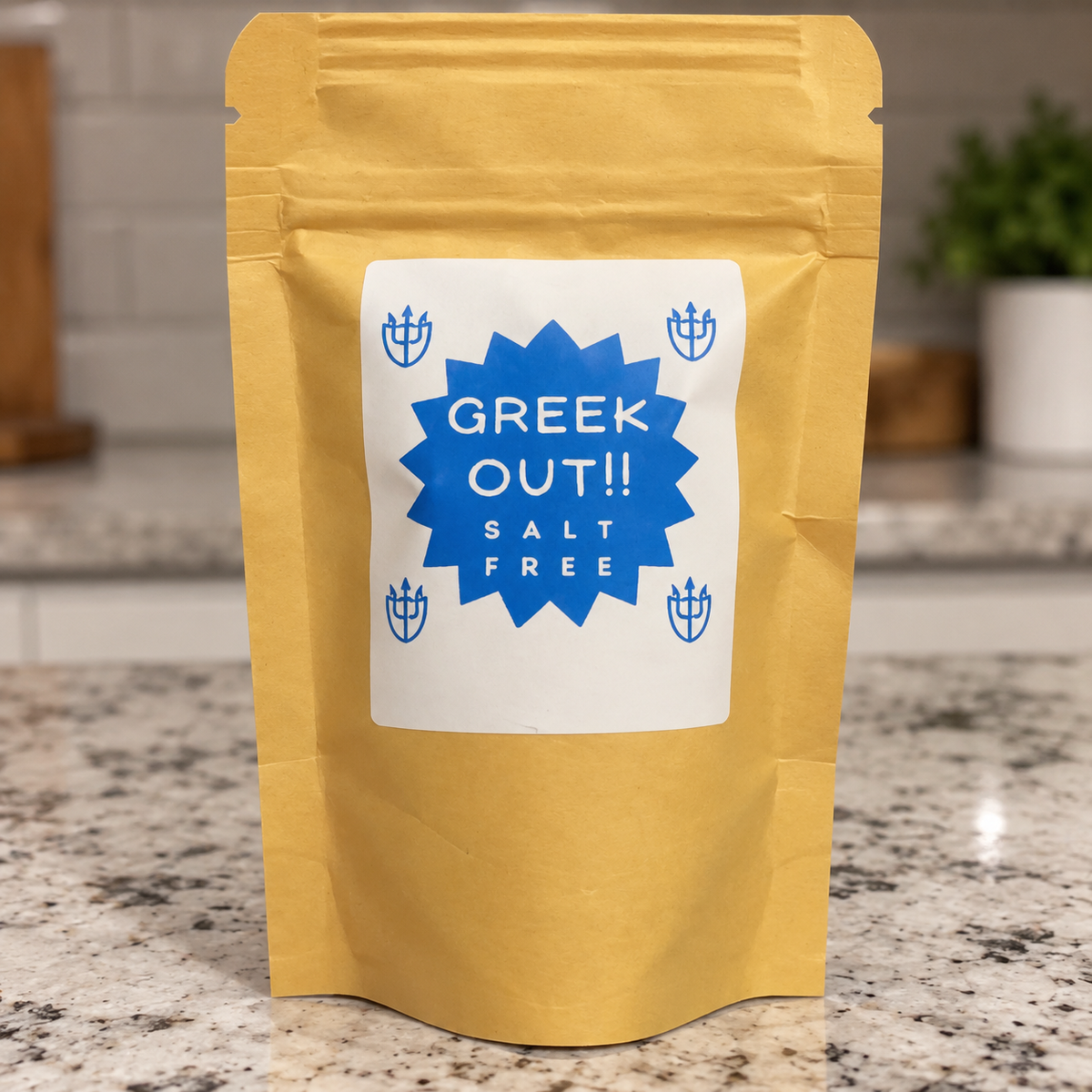 A stand-up kraft paper pouch of Greek Out!! Salt Free seasoning standing on a granite countertop in a kitchen setting. The pouch is golden yellow with a large white label featuring a blue starburst and blue trident icons.