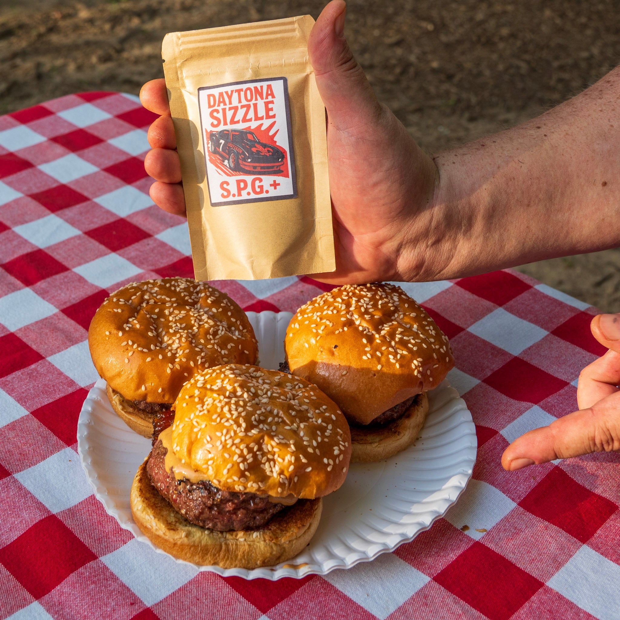 Hand holding a bag of Daytona Sizzle seasoning with three burgers on a plate on a red and white picnic tablecloth