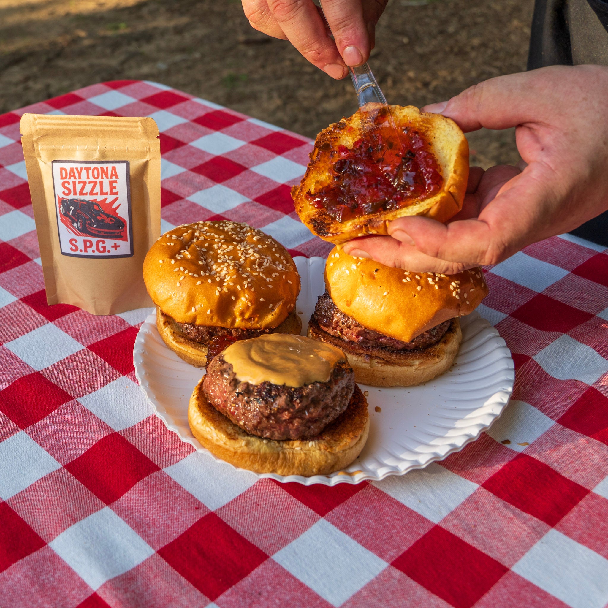 Hand spreading bacon pepper jelly on a burger bun with a bag of Daytona Sizzle and three burgers on a red and white picnic tablecloth