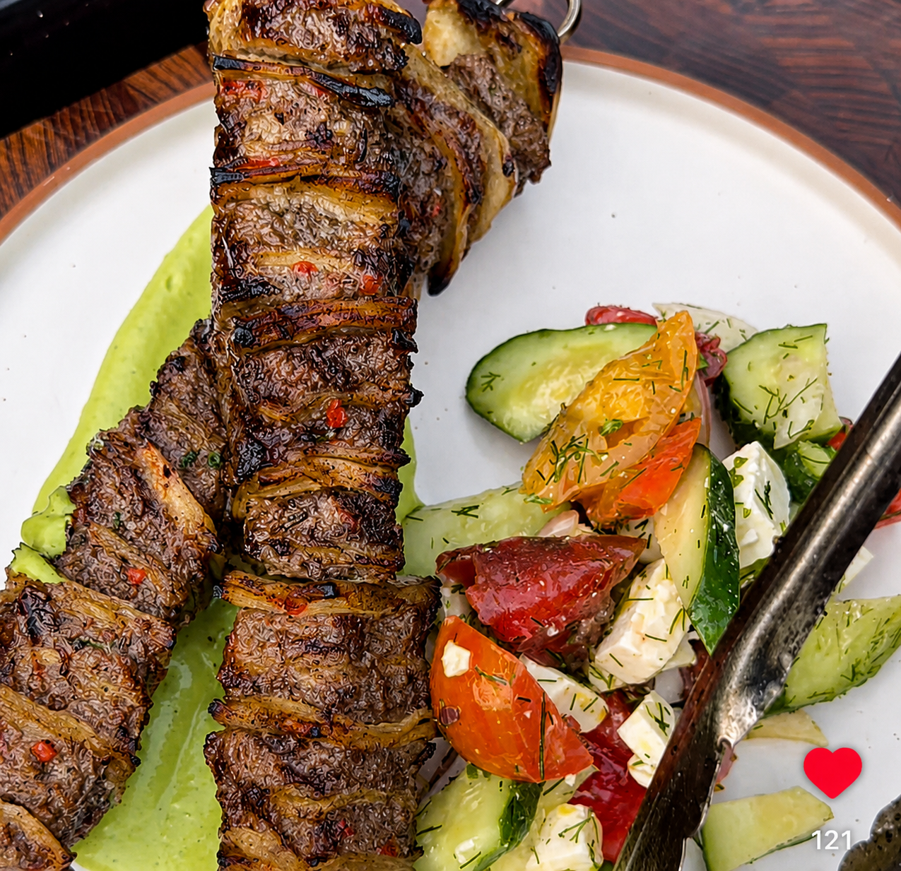 Grilled meat skewers served on a white plate with a side salad of tomatoes, cucumbers, and herbs, with a metal serving utensil visible. Greek Out Seasoning
