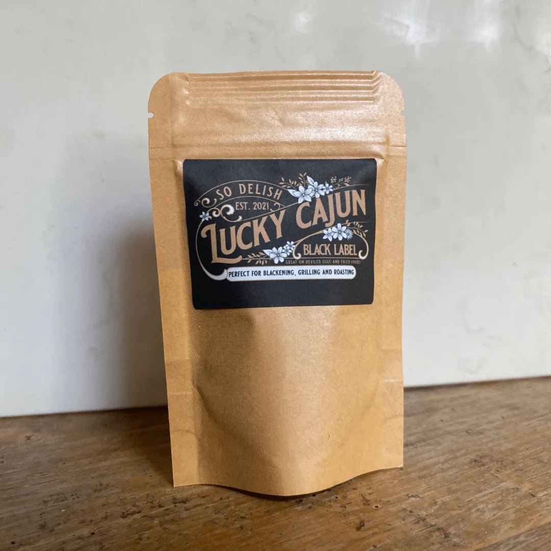 Lucky Cajun Black Label Authentic Cajun Seasoning pouch on a wooden table with a white background. Fresh-ground, small-batch Cajun rub featuring a Born On Date and no fillers.
