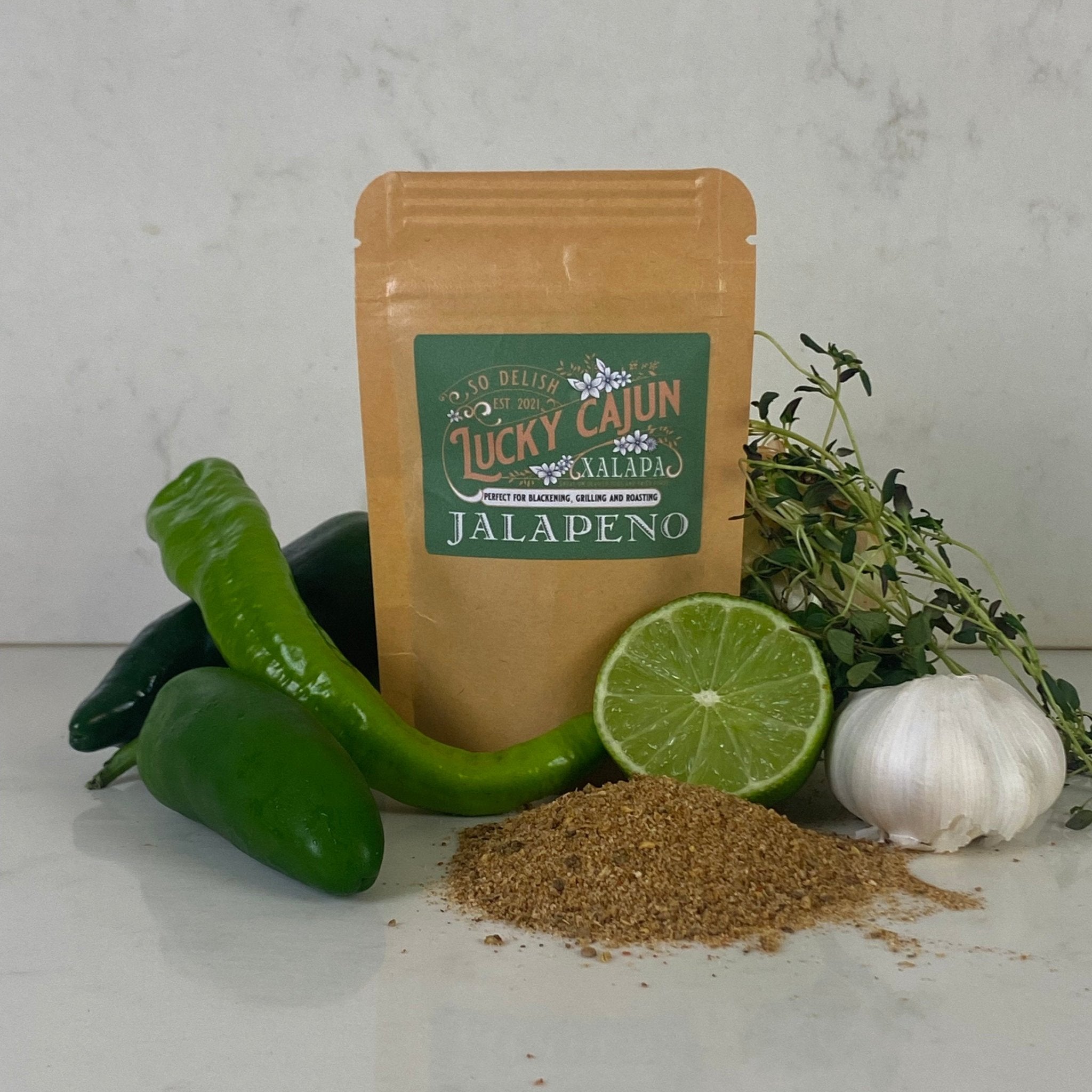 A pouch of Lucky Cajun Xalapa Jalapeño Seasoning surrounded by the fresh ingredients that define its flavor profile: whole jalapeños, garlic, lime, and aromatic herbs—the antidote to grocery store flavor sands.