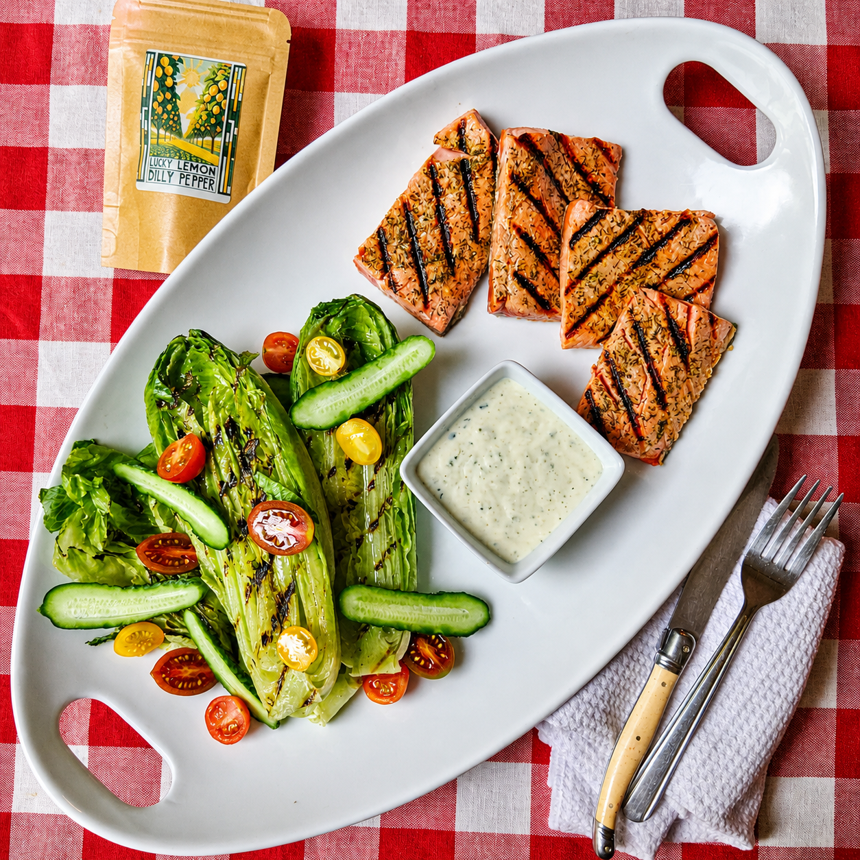 Grilled salmon fillets with Lucky Cajun Lemon Dilly Pepper seasoning served over smoky grilled romaine with sliced tomato cucumber and lemon caper herb dressing on a platter being served family style