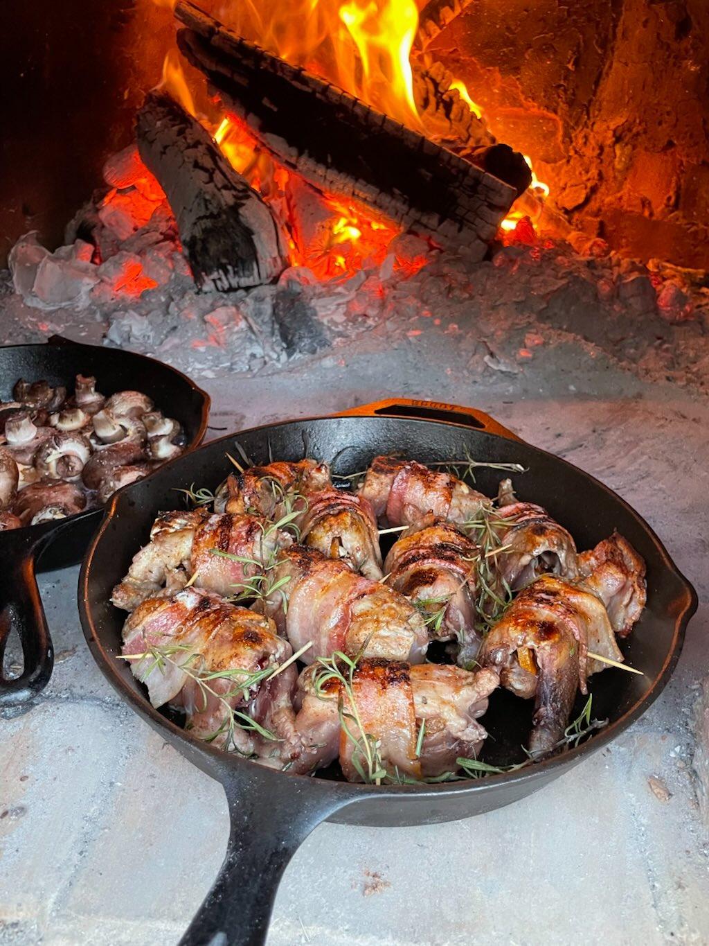 Bacon-wrapped quail in a cast iron skillet in a wood-fired oven, topped with fresh rosemary sprigs, stuffed with dates and pecans and seasoned with Lucky Cajun Black Label.