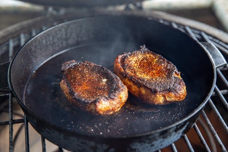 Two thick-cut blackened pork chops searing in a cast iron skillet over an open flame, developing a deep Cajun crust with Lucky Cajun Black Label seasoning.