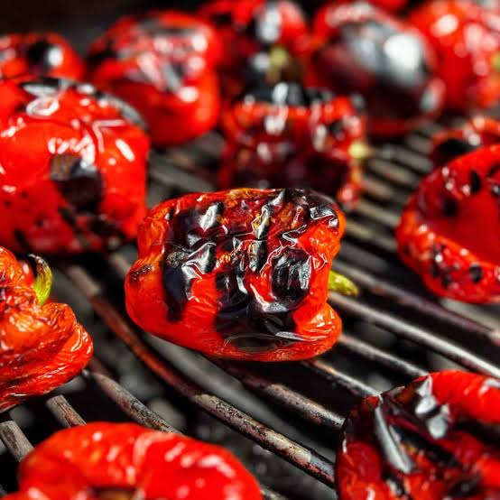 Red bell peppers charring on a grill showing the complete blackening needed to develop the smoky sweet flesh used in homemade romesco sauce