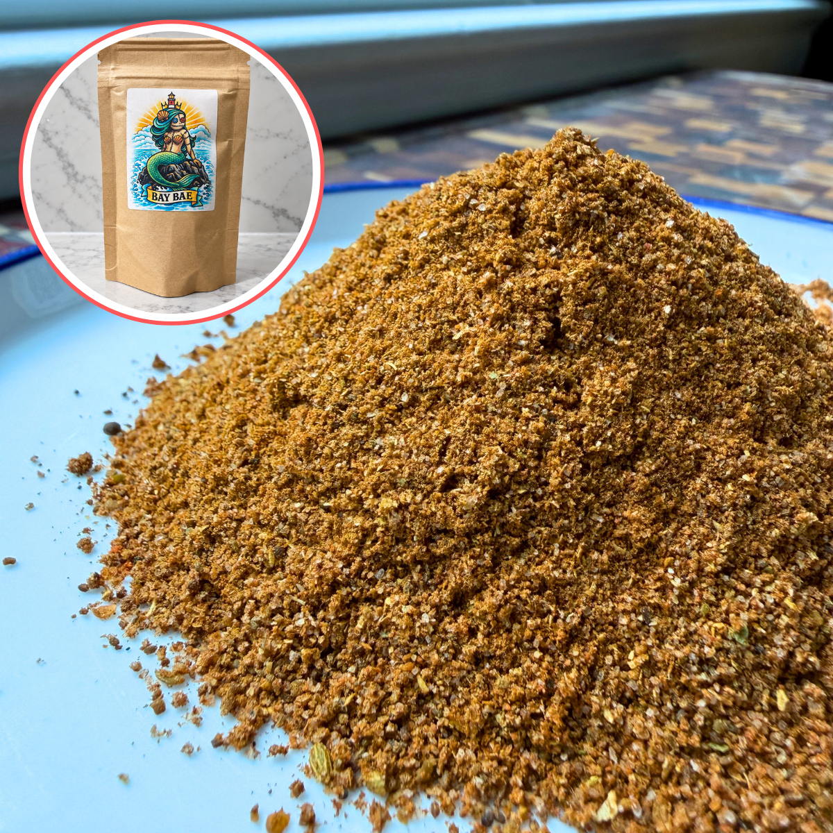 A pile of coarse spice blend on a light blue plate, with an inset showing a kraft paper pouch labeled 'Bay Bae' in the top-left corner.