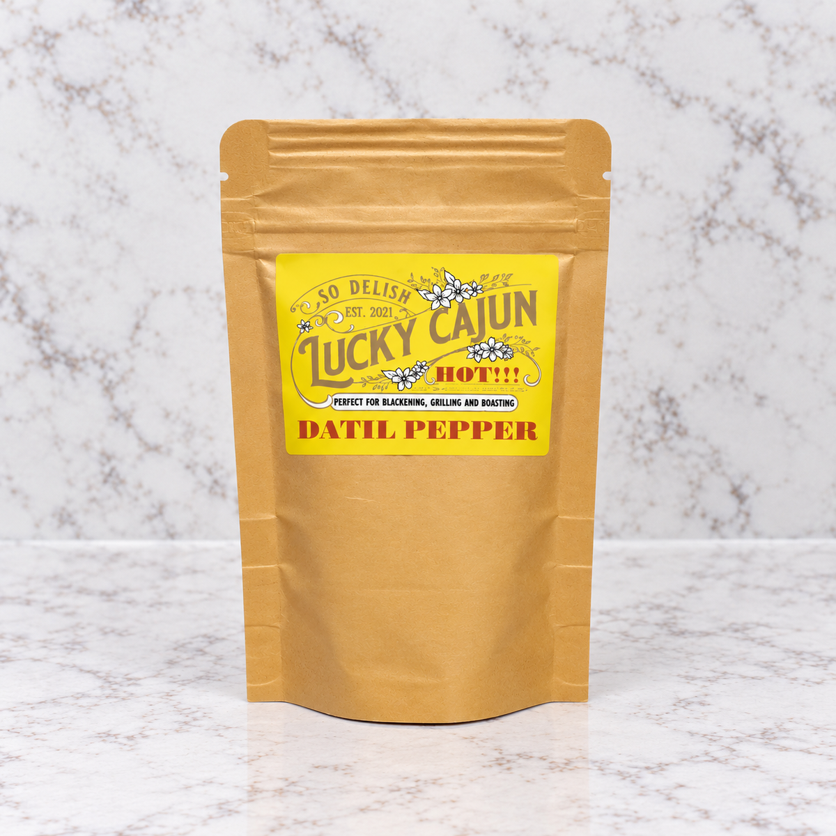 A stand-up kraft paper pouch of Lucky Cajun Datil Pepper seasoning, featuring a bright yellow label with white and red text, set against a white marble background.