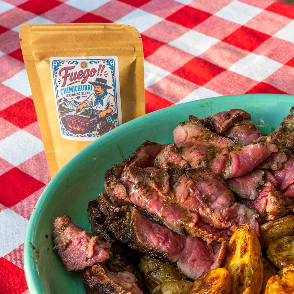 A pouch of Fuego!! Chimichurri seasoning sauce sits behind a large bowl of cooked sliced lamb and roasted potatoes on a red-and-white checkered tablecloth.