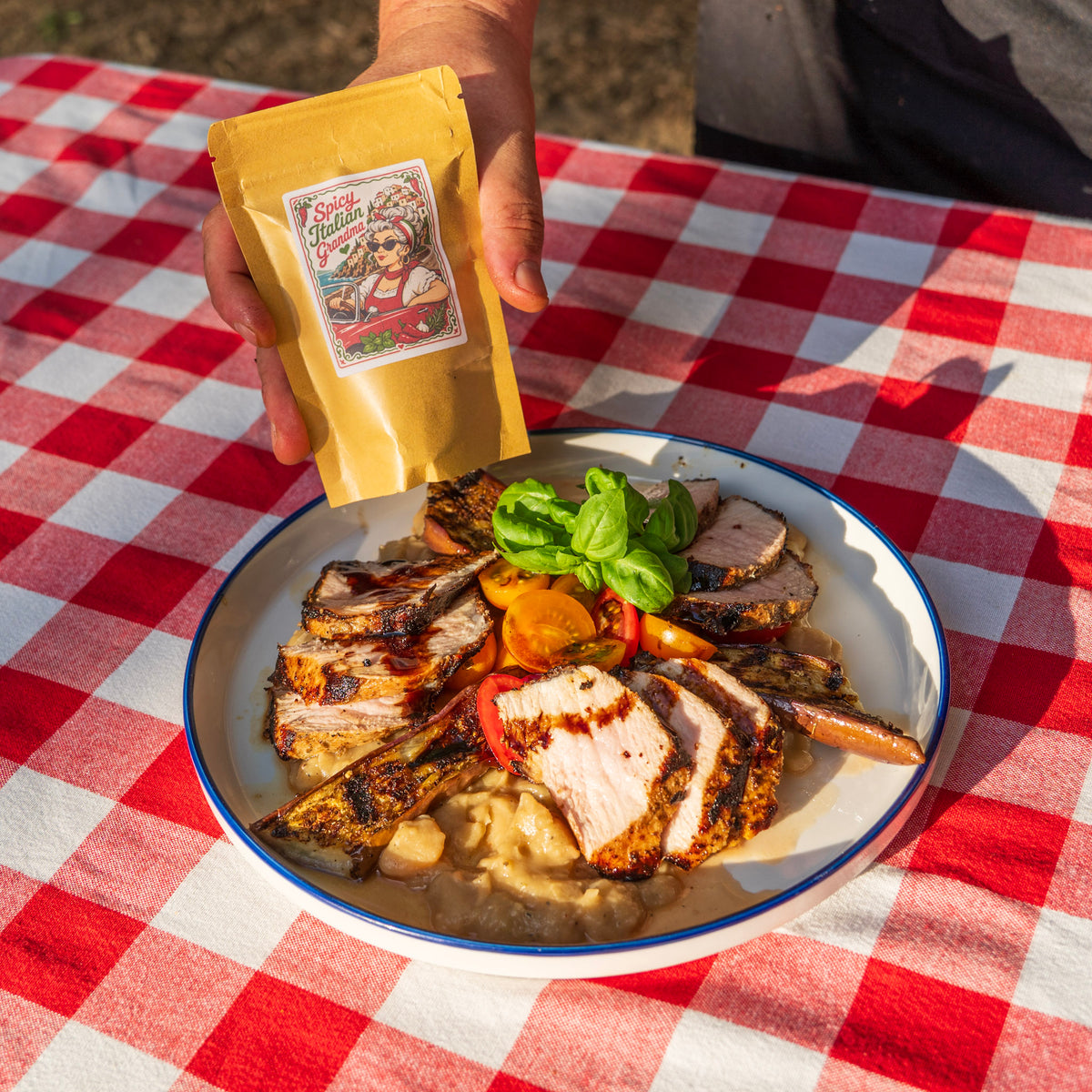 Hand holding a bag of fresh-ground Spicy Italian Grandma seasoning over a picnic table with sliced grilled pork loin, stewed eggplant and lima beans, topped with fresh tomato, basil, and a balsamic drizzle