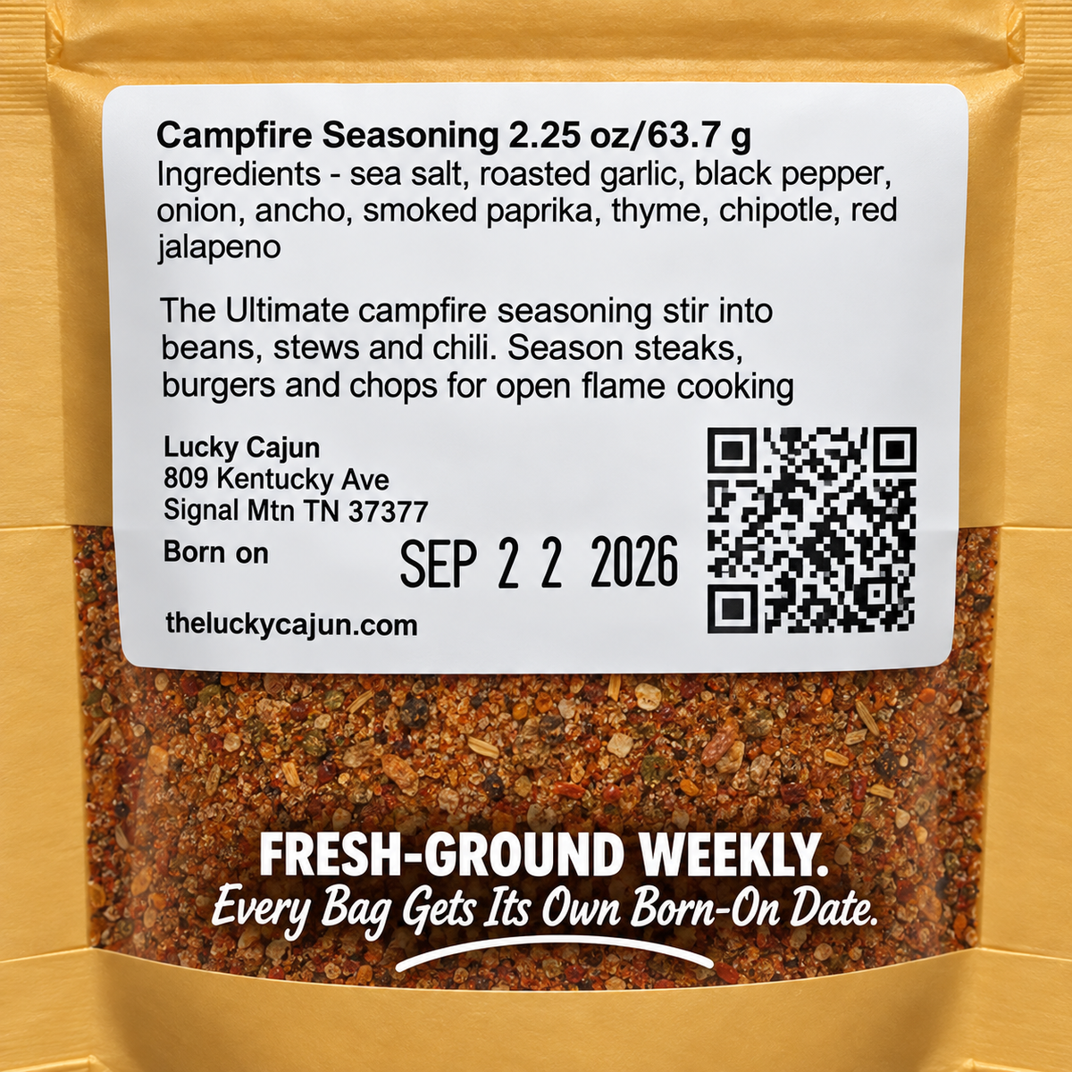 A bag of Lucky Cajun Campfire Seasoning with a large white label showing the ingredients, address, birth date, and QR code, set against a gold foil pouch with visible ground seasoning through the clear lower section.