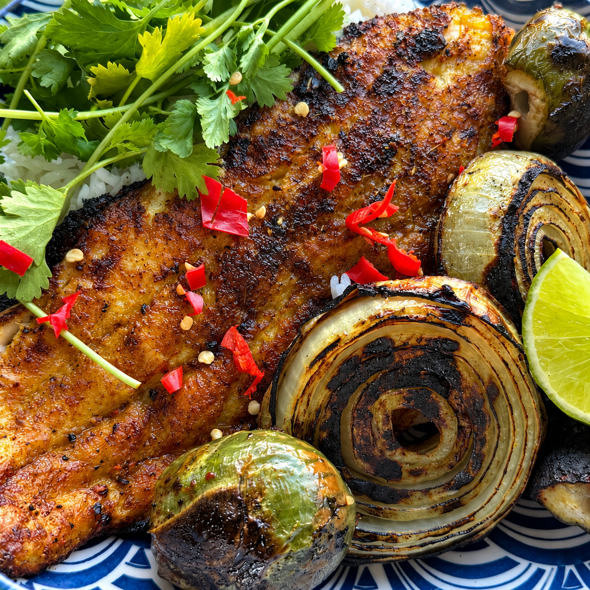 Grilled fish fillet served on a plate with grilled onions, Thai Eggplant, cilantro, rice, and lime wedges.