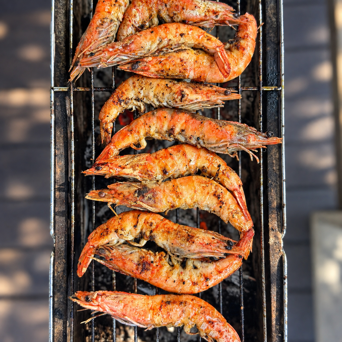 Grilled prawns cooking on a black metal grill outdoors, with visible char marks and seasoning on a dark background.