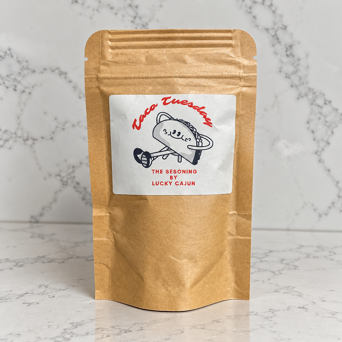 A kraft paper stand-up pouch with a white label reading 'Taco Tuesday' and 'The Seasoning by Lucky Cajun,' photographed on a light marble surface against a marble wall.