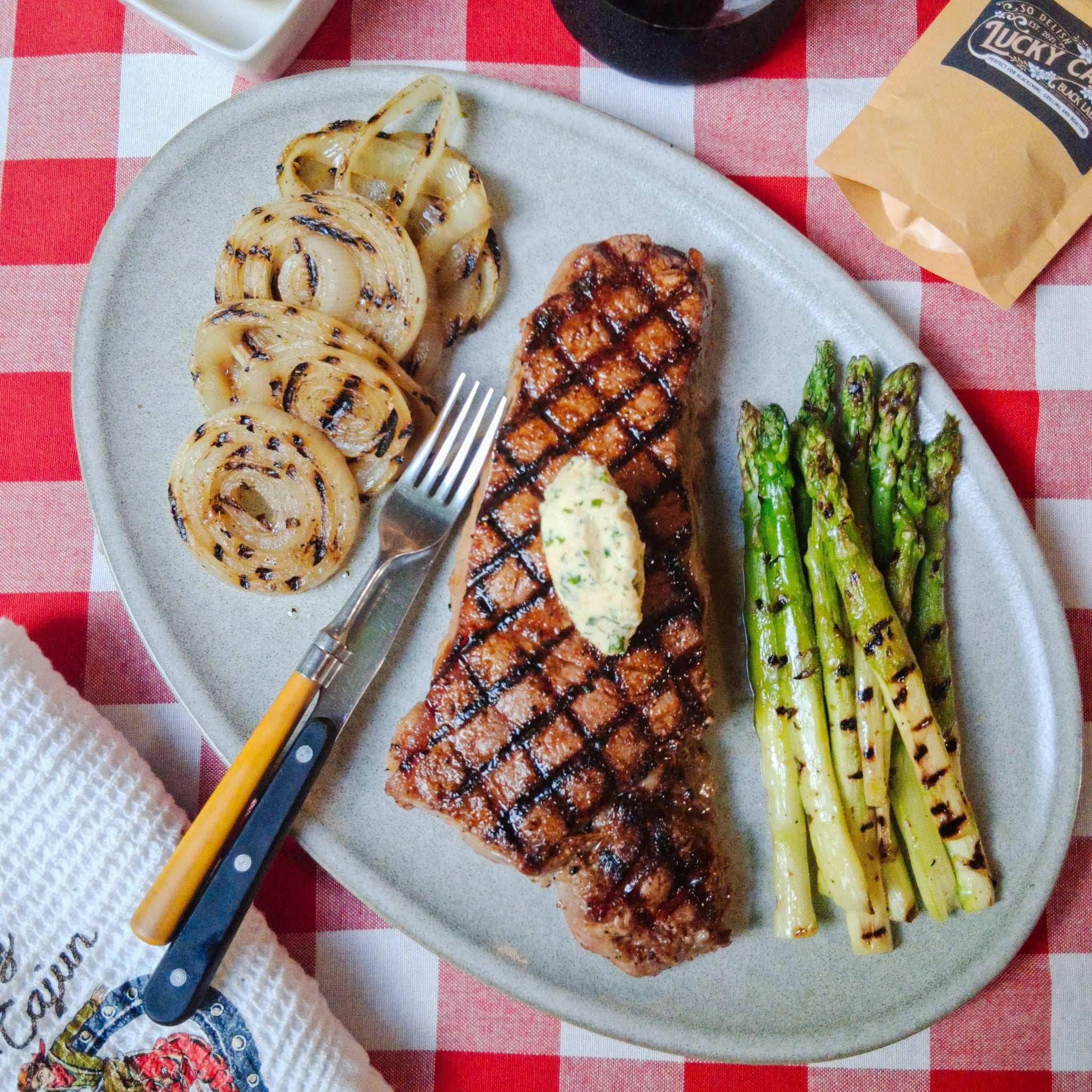 Grilled NY Strip steak seasoned with Lucky Cajun Black Label, topped with herb compound butter, and served with grilled asparagus and onions. Premium fresh-ground steak rub.