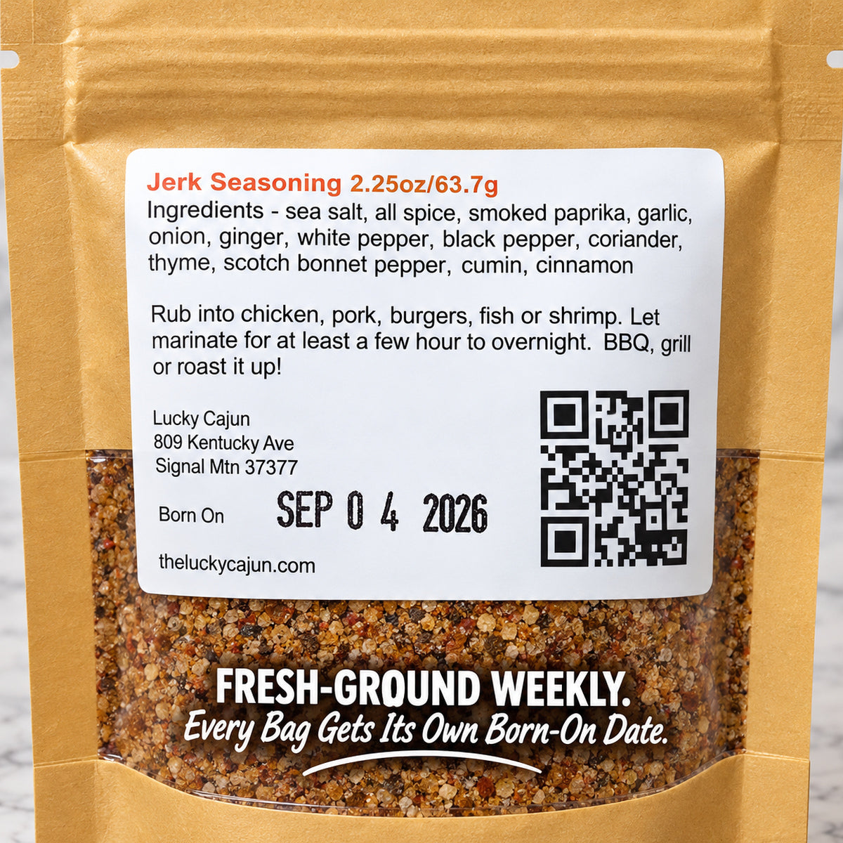 A bag of Lucky Cajun Jerk Seasoning with a large white ingredient label on the back, showing the product name, ingredients list, address, QR code, and 'Fresh-Ground Weekly' text. The seasoning granules are visible through a clear window in the kraft paper bag.