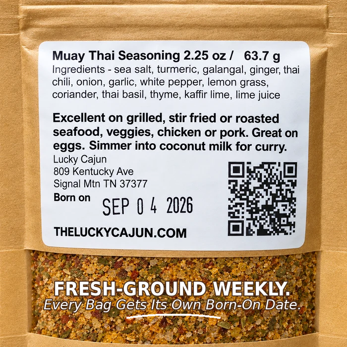 A kraft paper bag of Muay Thai Seasoning with a large white label showing ingredients, address, birth date, and a QR code. A clear window at the bottom reveals the fresh ground seasoning blend inside.